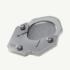 A brushed silver metal mechanical component, likely a watch movement bridge or clutch plate, featuring three hex screws and a small engraved logo, shown on a light off-white background.