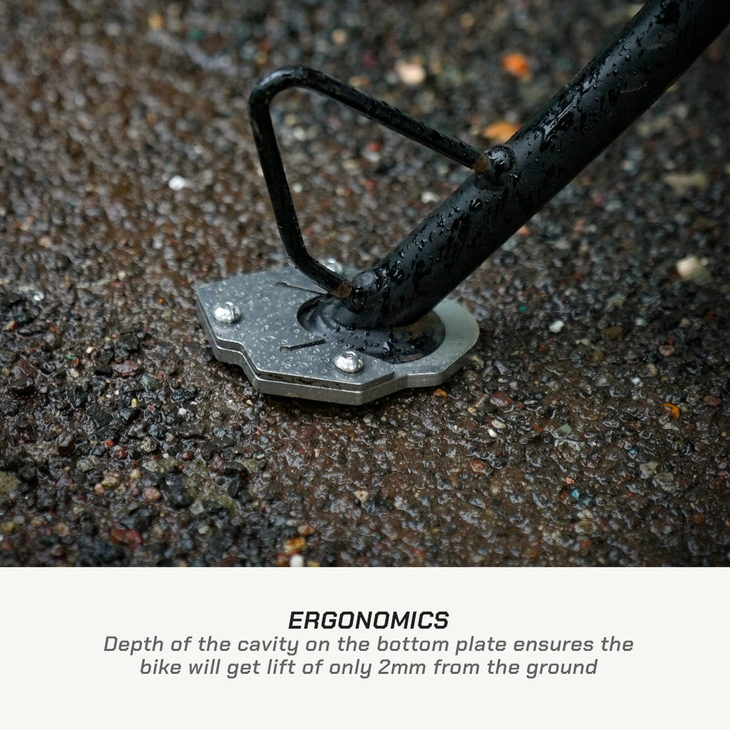 Close-up of a bicycle bottom bracket and pedal spindle mounted on wet asphalt, with text below reading 'ERGONOMICS' and a description about the bike lifting only 2mm from the ground.