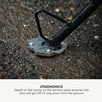 Close-up of a bicycle bottom bracket and pedal spindle mounted on wet asphalt, with text below reading 'ERGONOMICS' and a description about the bike lifting only 2mm from the ground.