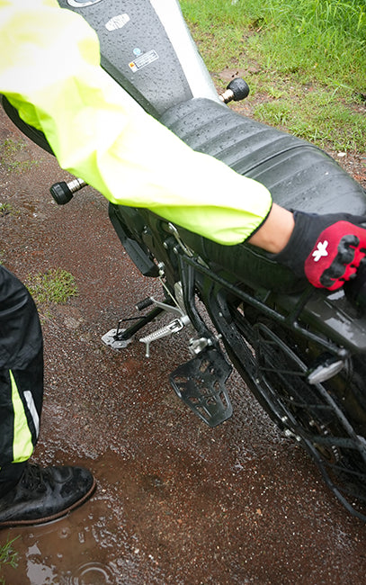 Specially designed to work on uneven surfaces A close-up outdoor shot of a person in a neon yellow rain jacket and black gloves touching a wet black motorcycle seat on a muddy, rainy day.