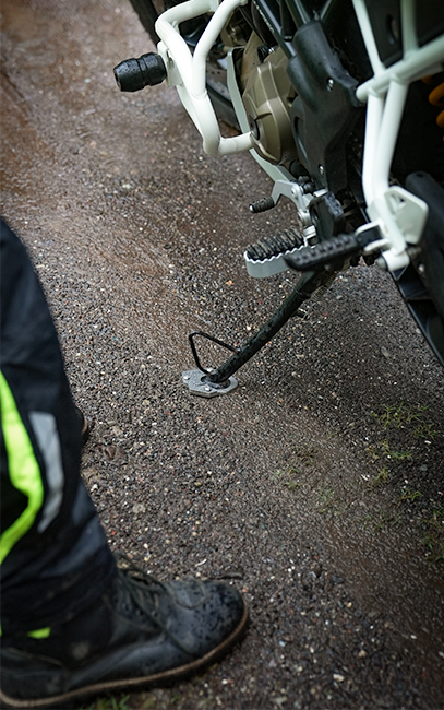 Better weight distribution Close-up of a motorcycle kickstand extended on wet gravel, with part of a person's black boot and dark pants with a bright yellow-green stripe visible at the left edge.