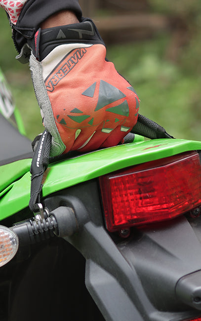 Comfort grip A close-up of a person's gloved hand resting on the green fender of a motorcycle, with the glove showing a bright orange, gray, and black design and the brand name 'MCI JERZA' visible.