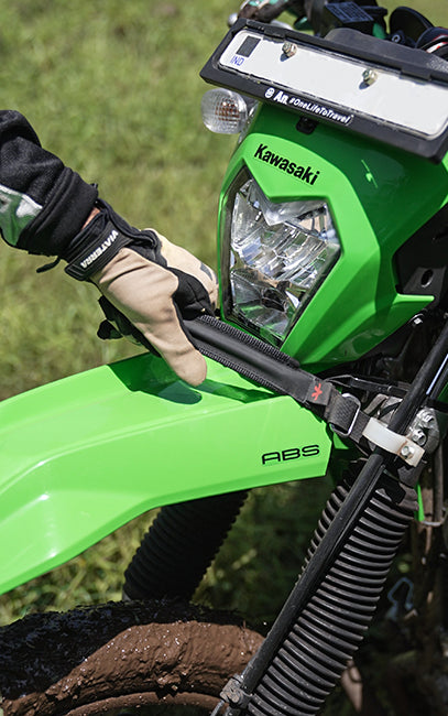 Comfort grip A close-up outdoor shot of a bright green Kawasaki ABS dirt bike, showing a person's gloved hand adjusting a black handlebar clamp or strap near the headlight and license plate area.