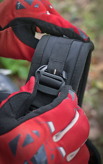DASH System (Dual Adjustable Sliding Harness) Close-up of a red and black glove holding a black adjustable strap with a plastic buckle, set against a blurred outdoor background.