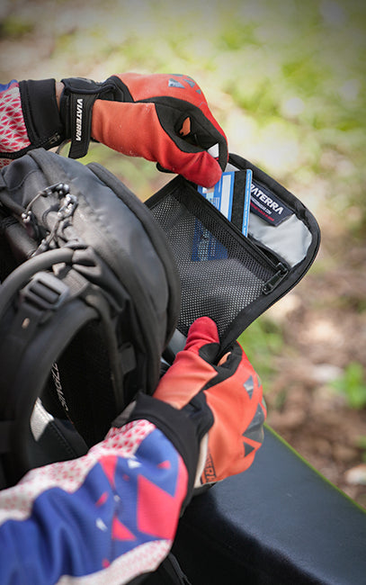 Dedicated Front Compartment A close-up of a person wearing orange and black Verva gloves, opening a black mesh card holder pouch inside a black backpack outdoors.
