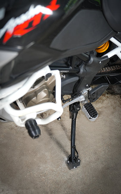 Designed_for TVS Apache RTX 300  Close-up of a motorcycle's rear suspension and kickstand, showing a black metal kickstand attached to a black and white motorcycle body with a visible orange rear shock absorber on a concrete floor.