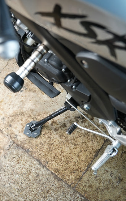 Designed for Yamaha XSR155 Close-up of a motorcycle's rear section showing the black engine casing, silver foot pegs, and black kickstand on a beige stone tiled floor.