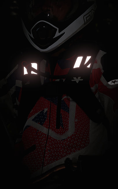 Enhanced Visibility A close-up, low-light shot of a person wearing a red, white, and black patterned motocross or snowmobile jacket with high-visibility reflective strips across the chest, along with a white and black helmet.