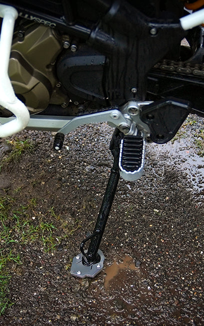 Ergonomics Close-up of a motorcycle's black and silver footpeg and kickstand assembly, shown outdoors on wet gravel and mud with part of the bike's engine visible.