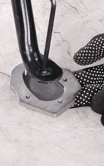 Better weight distribution A close-up of a black metal stand or frame component resting on a silver metal base, with a black-and-white dotted grip glove visible on the right, set against a light gray textured surface.