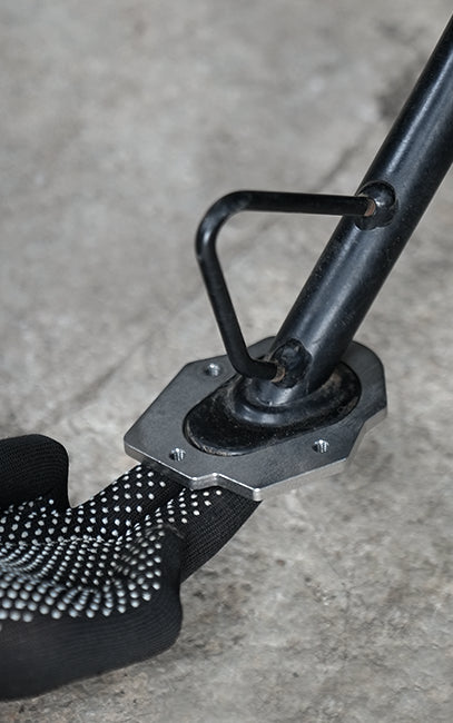 TVS Apache RTX 300 SIDESTAND SHOE Close-up of a black padded strap with a textured dotted grip and a metal mounting bracket attached to a black pole, set against a gray concrete floor.