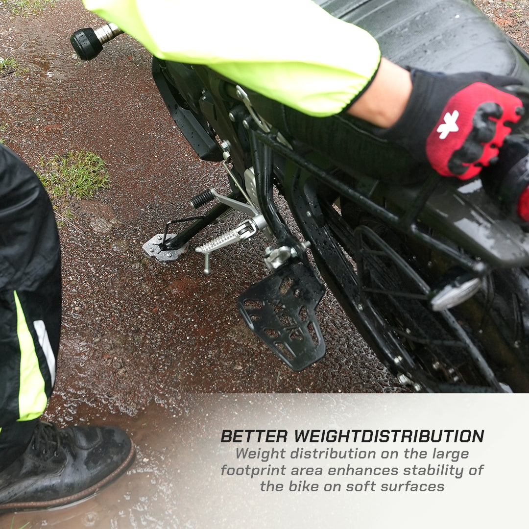 Close-up of a motorcycle's rear wheel and foot pegs with a rider's gloved foot resting on the pegs, outdoors on wet pavement. Text at the bottom reads: 'BETTER WEIGHTDISTRIBUTION Weight distribution on the large footprint area enhances stability of the bike on soft surfaces.