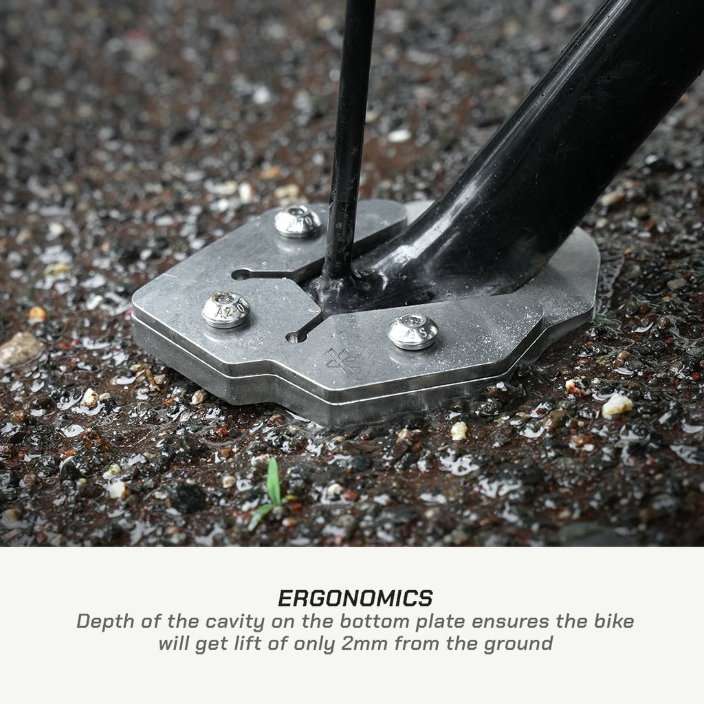 Close-up of a bicycle fork base mounted on a metal ground plate with three silver screws, set in gravel. Text below reads 'ERGONOMICS' with a description about the 2mm ground lift.