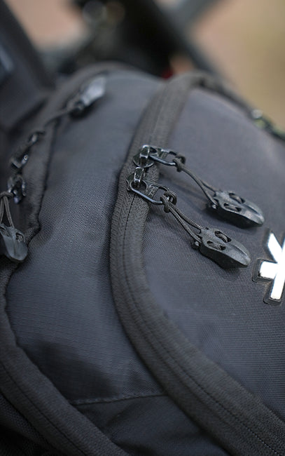 Genuine YKK Zippers Close-up of a dark gray fabric bag with black zipper pulls and a white stylized cross logo on the front.