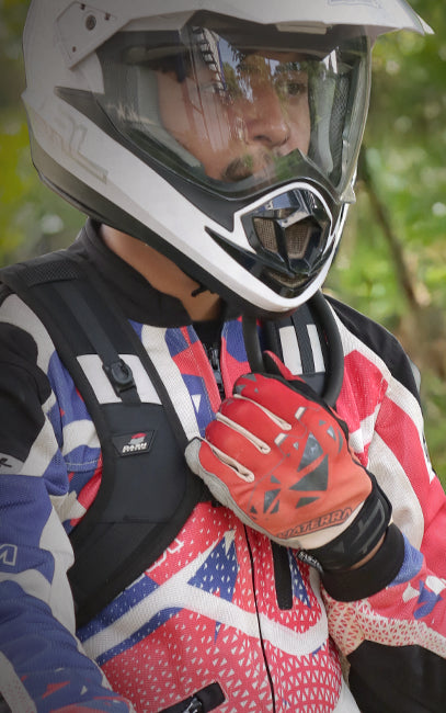 Hydrated, Focused A close-up of a person wearing a white full-face motorcycle helmet with a clear visor, a red, white, and blue patterned riding jacket, a black backpack, and a red glove.
