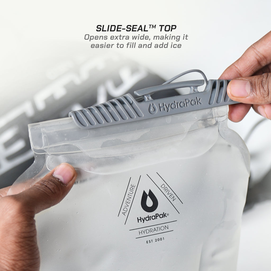 A clear HydroPak hydration pouch being held by two hands, with a gray slide-seal closure at the top. Text at the top reads 'SLIDE-SEAL TOP' and 'Opens extra wide, making it easier to fill and add ice.