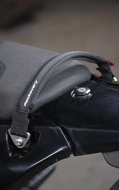Improved ergonomics Close-up of a black motorcycle seat bag with a textured fabric body and a black strap secured to the bike frame, featuring the brand name 'RetroRevvy' printed on the side.