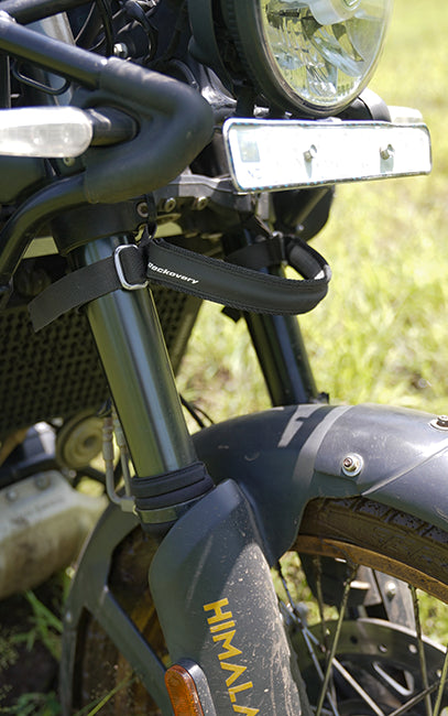 Improved ergonomics Close-up of a black motorcycle front suspension strap with a buckle, attached to the fork, on a motorcycle outdoors with grass in the background.