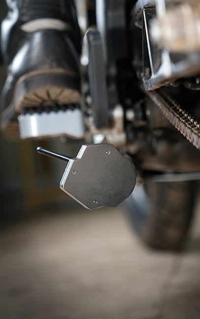 Longer thread life A close-up shot of a motorcycle's foot peg and chain guard, with a foot resting on the peg. The chain guard is a dark gray, octagonal metal plate with a small metal tool inserted into its center.