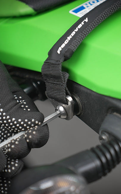 Improved ergonomics Close-up of a gloved hand tightening a metal bolt on a black recovery strap attached to a bright green equipment bag. The strap has the word 'recovery' printed in white.