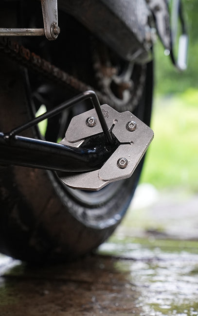 Precision & Consistency Close-up of a motorcycle or scooter rear wheel with a black metal kickstand attached to a silver metal mounting plate, set outdoors on wet pavement with blurred green foliage in the background.
