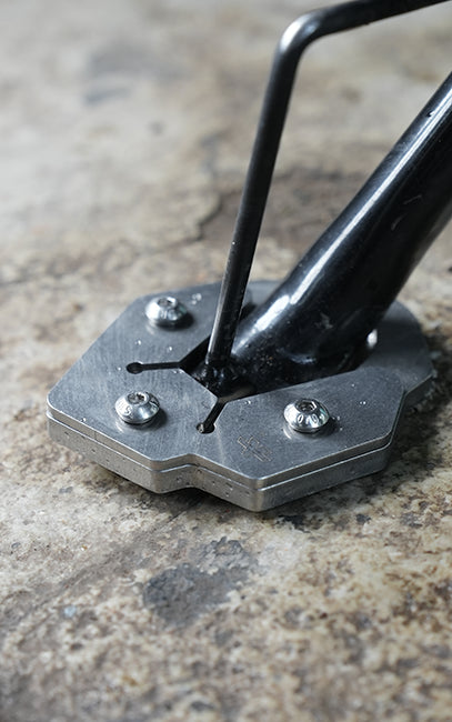 Premium build and finish Close-up of a metal bicycle fork base clamp with three silver screws, shown installed on a textured concrete surface.