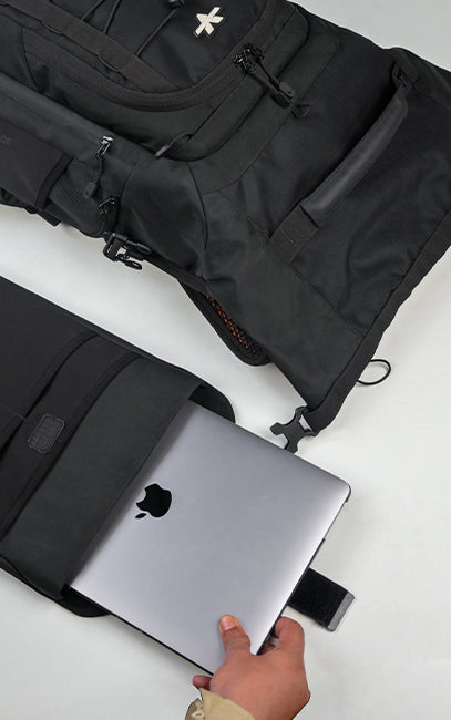 Modular Laptop Sleeve A black laptop sleeve with a silver Apple MacBook inside, held by a hand on a white surface, with a larger black backpack shown in the background.