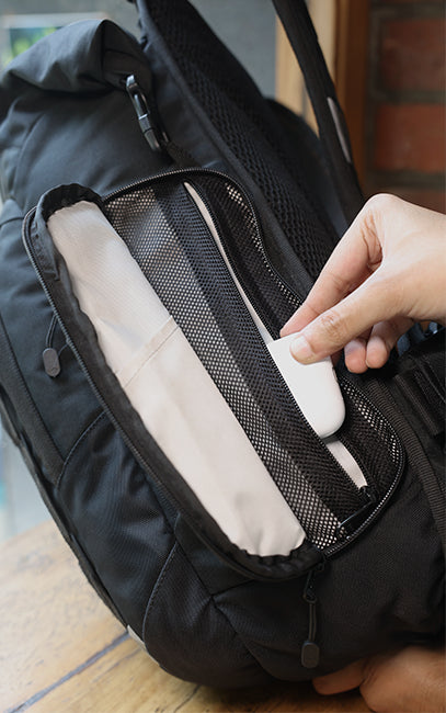 Organized Side Pocket A close-up of a black backpack with a hand placing a white rectangular item into the open front mesh pocket.