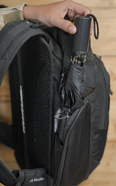 Utility Side Pocket A close-up of a black backpack with a side pocket holding a black umbrella, with a hand adjusting the umbrella handle. The brand name 'Skelle' is visible on a small tag near the bottom left.
