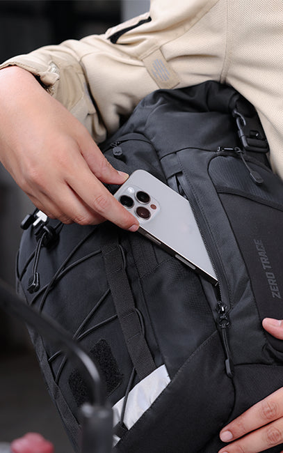 Quick-Access Waterproof Pocket A close-up of a person wearing a beige jacket and holding a black backpack with a silver smartphone tucked into a side pocket. The backpack has visible branding that reads 'ZERO TRACE'.