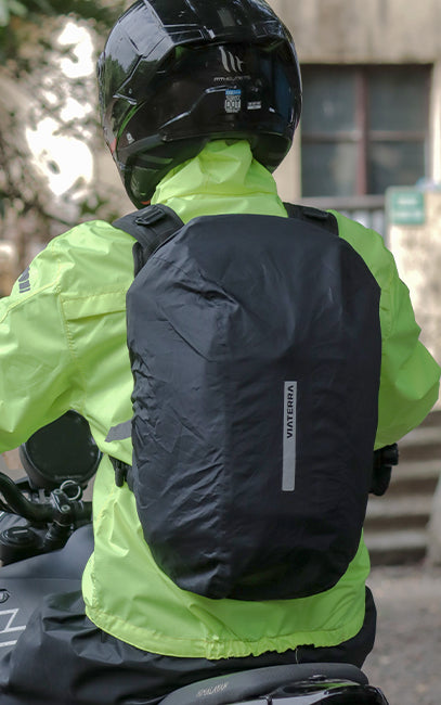 Integrated Rain Cover A person wearing a black motorcycle helmet and a high-visibility neon yellow-green jacket, seen from behind while riding a motorcycle. They are wearing a black waterproof backpack cover with a vertical white brand label that reads 'VANGUARD'.