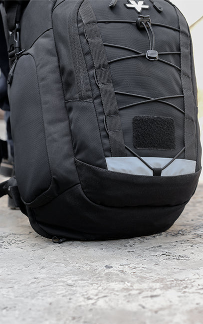 Reinforced Base A close-up of a black tactical backpack with crisscross compression straps and a reflective gray panel near the bottom, resting on a concrete surface.