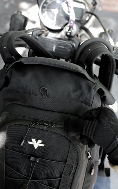 Durable Zippers A close-up of a black tactical motorcycle backpack mounted on a handlebar, with a gloved hand adjusting the front strap. The backpack features a white stylized 'V' logo on the front pocket.
