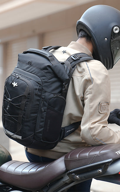 ASM (Adaptive Suspension Module) A black motorcycle backpack with multiple compartments and a white logo, worn by a rider in a beige jacket and black helmet sitting on a brown motorcycle.