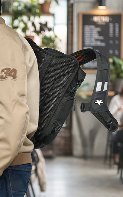 Ventilated Harness & Back Panel A black mesh and fabric crossbody sling backpack worn by a person in a cafe setting. The backpack features a prominent black mesh back panel, a zippered front pocket, and a black shoulder strap with white reflective stripes and a white logo.
