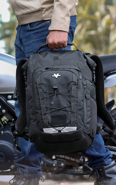 Form-Stable Handle A black tactical-style backpack with a top handle and front zippered compartments, held by a person wearing blue jeans and a beige jacket next to a motorcycle.