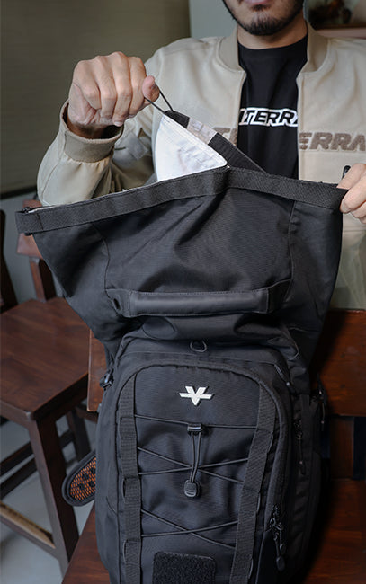 Removable Waterproof Liner A black tactical backpack with a white stylized logo on the front pocket, being held open by a person wearing a beige jacket and black t-shirt.