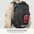 A black backpack shown on a model from the back, featuring a front pocket with an elastic bungee cord holding a red and black glove. The model is wearing a beige jacket against a light gray background, with text at the bottom explaining the EBC storage feature.