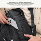Close-up of a black backpack with a quick-access waterproof pocket being used to store a silver smartphone. A person's hands are visible holding the phone and adjusting the strap, with a beige jacket in the background.