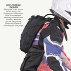 A black motorcycle backpack shown on a rider wearing a white helmet and a red, white, and blue riding jacket. The image includes text on the left explaining the low-profile design of the Singletrack backpack.