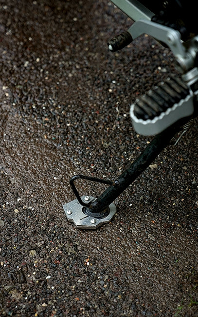 Specially designed to work on uneven surfaces Close-up of a motorcycle or scooter kickstand extended onto wet asphalt pavement, showing the metal foot peg and mounting plate.