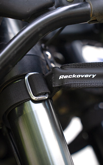 Strong Alloy Buckles Close-up of a black Reckovery motorcycle handlebar bar end strap secured around a metallic fork tube with a silver buckle.