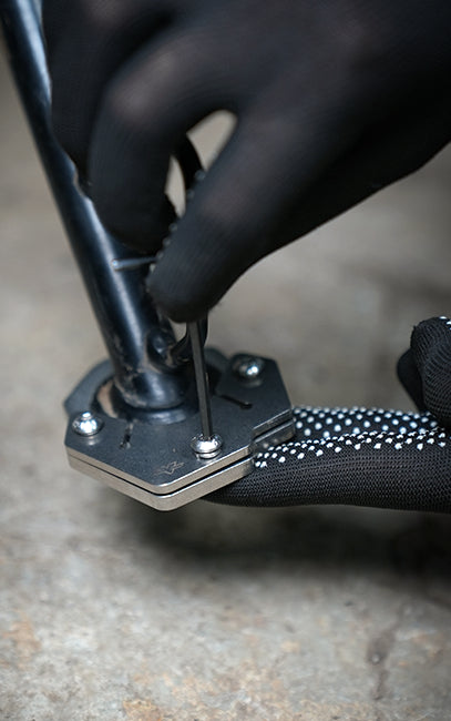 Strong clamping of shoe A close-up of gloved hands using a black-handled tool with a metal socket and hex key to adjust or tighten a bolt on a bicycle or similar component, set against a blurred concrete background.