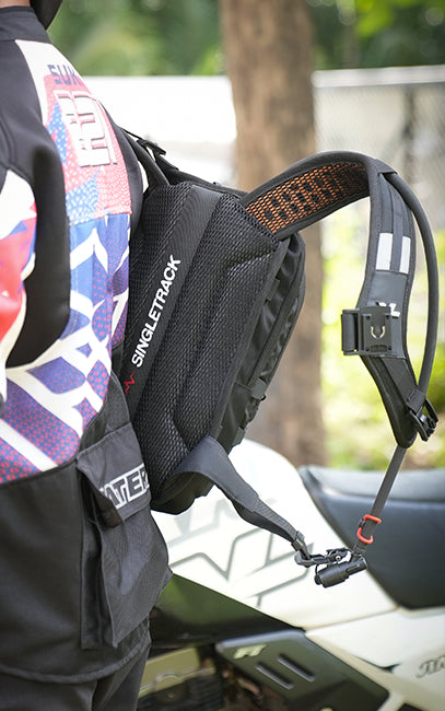 Ventilated Harness & Back Panel A close-up of a black motorcycle backpack with orange accents and reflective straps, worn by a rider in a patterned jacket next to a white motorcycle outdoors.